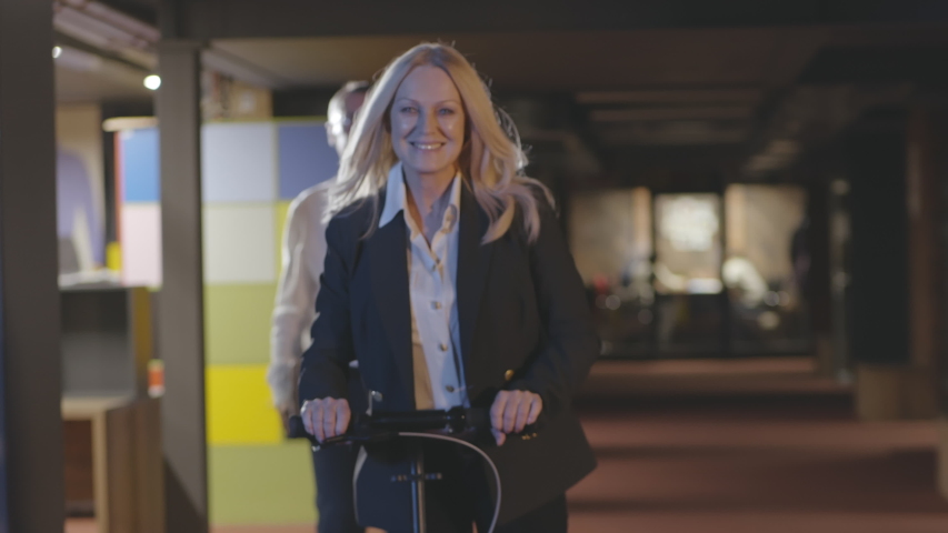 Business people talking and walking while their female colleague riding scooters over spacious corridor. Carefree mature employee in formal outfit riding kick scooter in hallway of office building