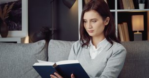 Charming young lady in domestic clothing sitting comfortably on couch and reading interesting book. Female person with dark hair enjoying literature at home.