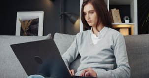 Concentrated young woman with dark hair sitting on grey couch and using personal laptop. Beautiful female in casual clothing working remotely from home.