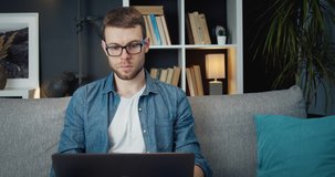 Bearded man in casual clothing working on laptop while sitting on couch at living room. Handsome male in eyeglasses using personal computer for work on distance.