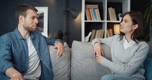 Happy young couple in domestic clothing sitting on grey couch and talking with each other. Man and woman spending free time together at home.