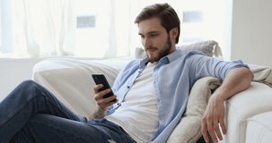 Happy millennial guy relaxing on sofa checking social media, playing mobile game or ordering food delivery holding using smart phone at home. Smiling man spending time at home with mobile gadget.
