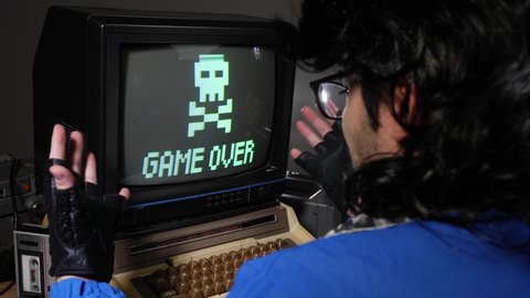 Upset, mad computer geek getting frustrated and hitting old concept computer from 70s 80s because of loosing and seeing game over sign
