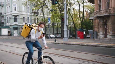 Poartrait of courier with yellow backpack and protective mask rides a bicycle on the street through the city with food delivery and using smartphone
