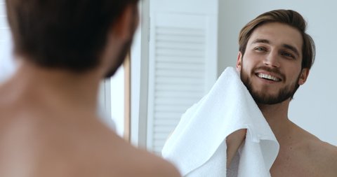 Smiling attractive man wiping face and beard holding towel looking in bathroom mirror. Happy handsome shirtless bearded guy washing face, shaving, grooming doing morning hygiene routine concept.