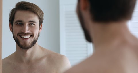 Funny confident young handsome man speaking to his reflection looking in bathroom mirror. Smiling sexy millennial guy narcissist touching beard feeling proud of himself shows good self-esteem concept.