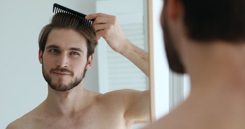 Handsome confident young man brushing hair holding comb looking in bathroom mirror. Attractive bearded shirtless guy doing hairstyle getting ready in the morning. Men haircare and styling concept.