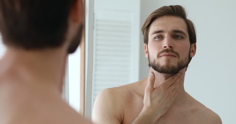 Confident bearded sexy shirtless man holding lotion bottle applying moisturizing balm on face and neck after shaving looking in bathroom mirror. Aftershave skin care treatment concept. Close up view