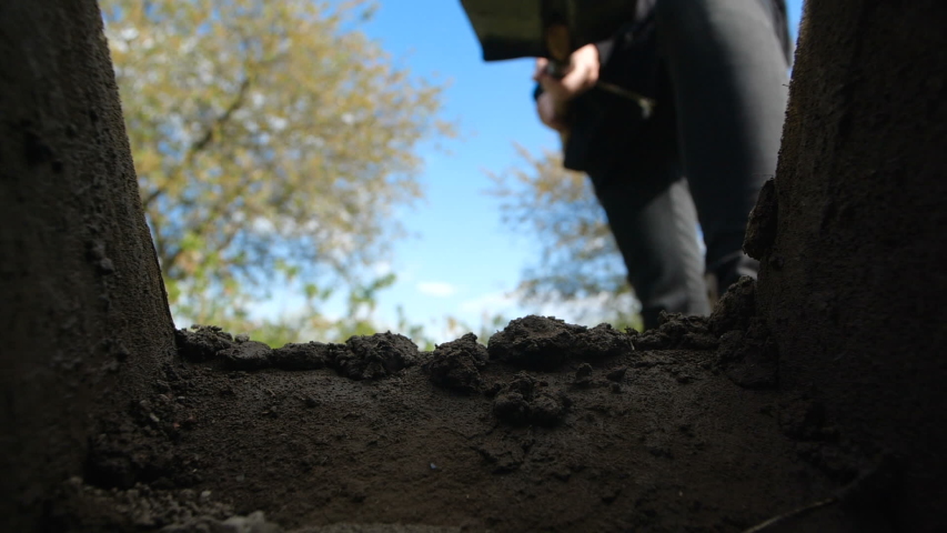 An unrecognizable person in dark clothes is digging a square pit against a blue sky with clouds. Point of view from the grave into which soil is sprinkled from a shovel. Slow motion Royalty-Free Stock Footage #1052102917