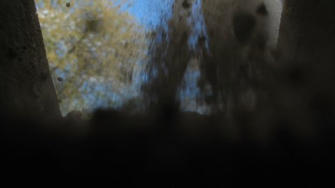 An unrecognizable person in dark clothes is digging a square pit against a blue sky with clouds. Point of view from the grave into which soil is sprinkled from a shovel. Slow motion