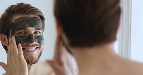 Smiling young bearded metrosexual man applying charcoal clay facial mask looking in bathroom mirror. Handsome shirtless guy cleaning face skin with spa cleansing mud. Male skincare beauty treatment.
