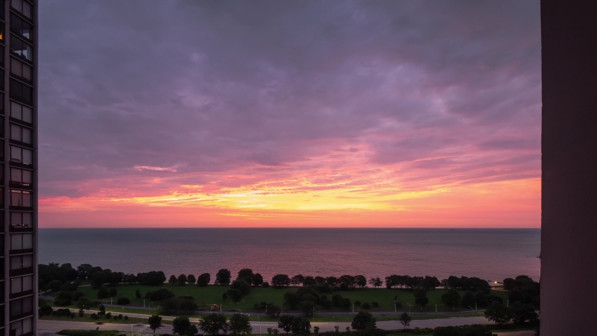 An incredible aerial timelapse of a yellow pink purple and blue sunrise reflects on the water of Lake Michigan as it rises over the horizon with traffic zooming by on Lake Shore Drive below.