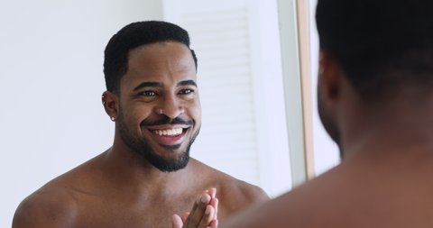 Handsome confident young adult afro american man looking in mirror. African sexy shirtless bearded millennial hipster guy enjoying grooming, getting ready, doing morning beauty routine in bathroom.