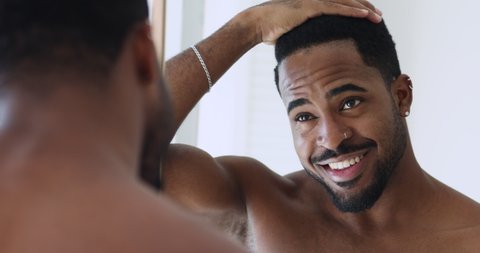Happy confident afro american hipster guy doing hairstyle looking in mirror. Smiling sexy bare african young man enjoying anti-dandruff shampoo hair result in the morning, grooming stand in bathroom.