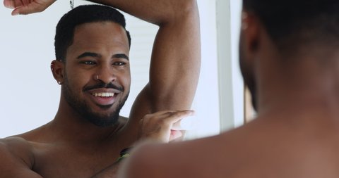Smiling sexy shirtless african american young man applying deodorant antiperspirant stick in armpit looking in bathroom mirror. Male daily hygiene hyperhidrosis treatment sweat protection concept.