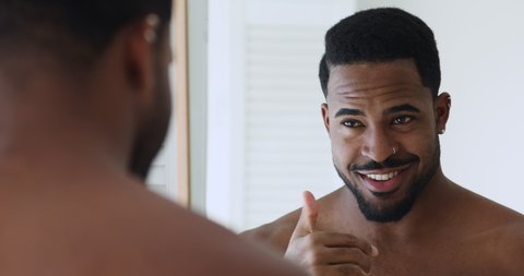 Confident young handsome african shirtless man pointing at bathroom mirror looking at own reflection. Smiling sexy afro american guy practicing in self confidence, grooming, getting ready in morning.