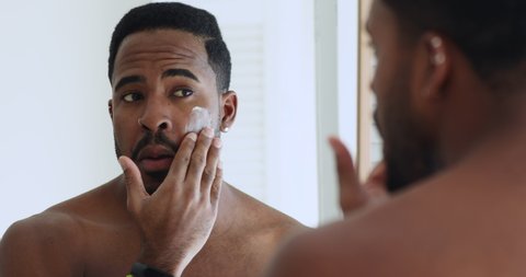 Young adult african metrosexual man applying moisturizer cream on face skin looking in bathroom mirror. Handsome bearded ethnic hipster guy puts facial creme on doing skincare morning beauty routine.