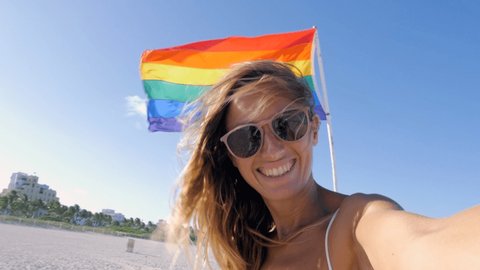 Gay woman taking selfie with rainbow flag on beach in Miami. LGBT concept people and sexuality choices. Human rights and respect - Slow motion