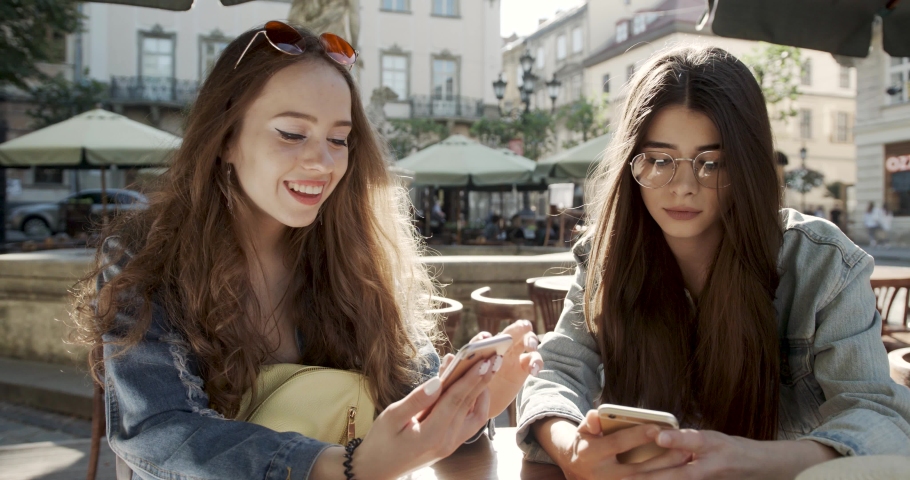 Two charming young girls using mobile phones while sitting at the cafe outdoors and pointing finger. Two Beautiful freelance woman use social media on smartphone in coffee shop, modern generation