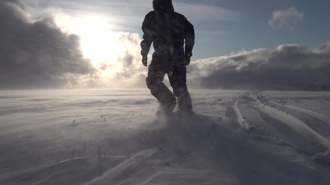 Silhouette of unidentified man in black snowmobile overall and helmet, he walking towards Sun shining through heavy clouds in mountains, poor snow visibility, high speed snow drift conditions.