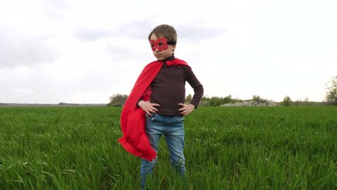 A happy child stands against the background of a green meadow, holding his hands on his belt and looking into the distance. A boy in a red Cape and mask imagines a superhero. Superman's red Cape