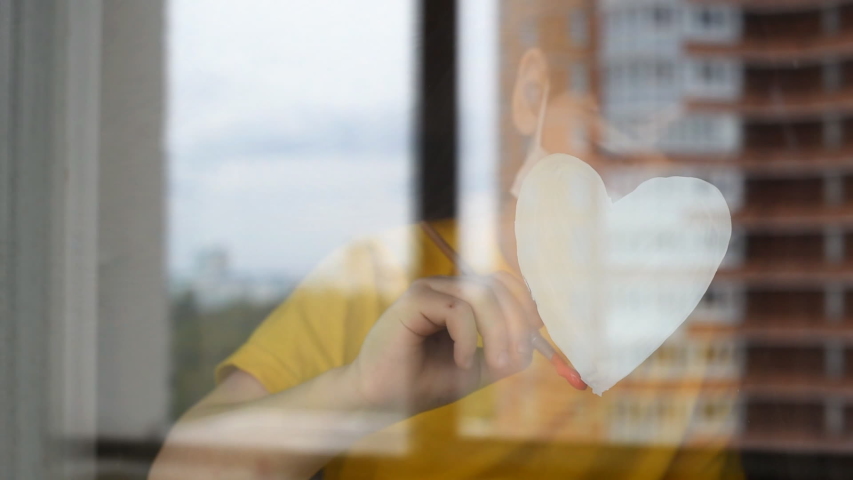 Child, little girl in a medical mask draws, paints a red heart simbol love, on the window glass. Self-isolation people, quarantined, stay at home, coronavirus pandemic