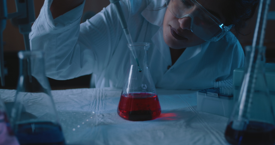Female research scientist adding a drop into a conical flask with red liquid.Blue lighting in a dark lab room.Medium, dolly out, slow motion, shot with BMPCC 4K.Concept: chemistry,science
