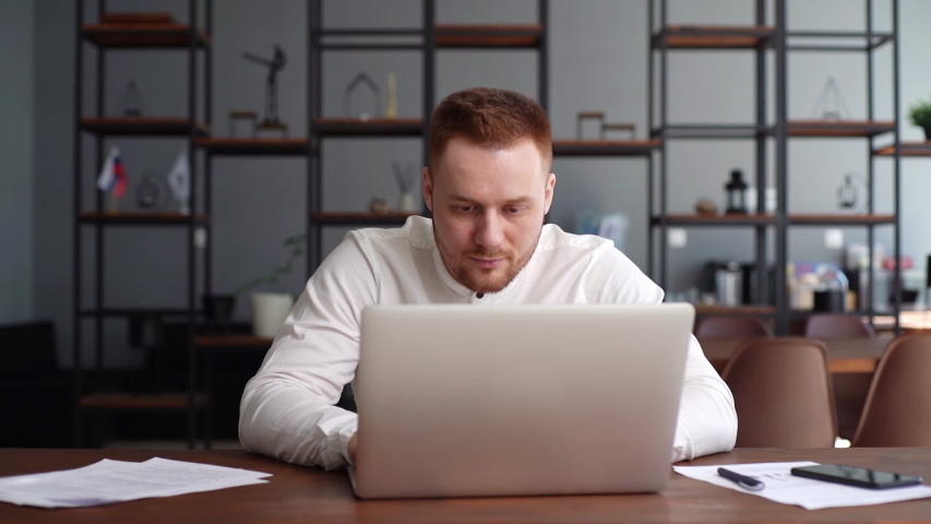Handsome bearded young businessman wearing fashion casual clothing is typing on laptop computer at the desk in modern office room. Concept of office working. Shotting in slow motion, | Shutterstock HD Video #1052148151