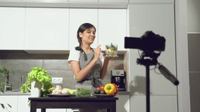 Attractive young woman recording a video about healthy eating on digital camera in the kitchen at home. Vlogging and social media concept