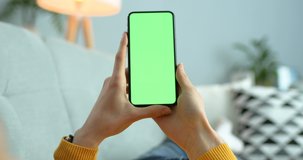Close up of vertical black smartphone with green screen in hands of Caucasian woman. Indoor. Mobile phone with chroma key. Place for ads. Female holding cellphone vertically in room.