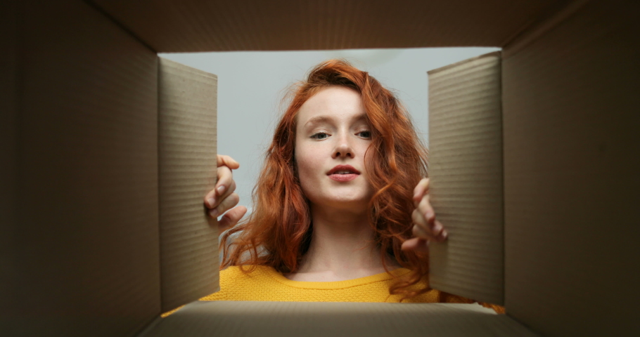 View from inside of cardbox on young cheerful Caucasian woman with red hair who looking in it and smiling. Close up of pretty happy smiled girl opening carton box. Female getting parcel.