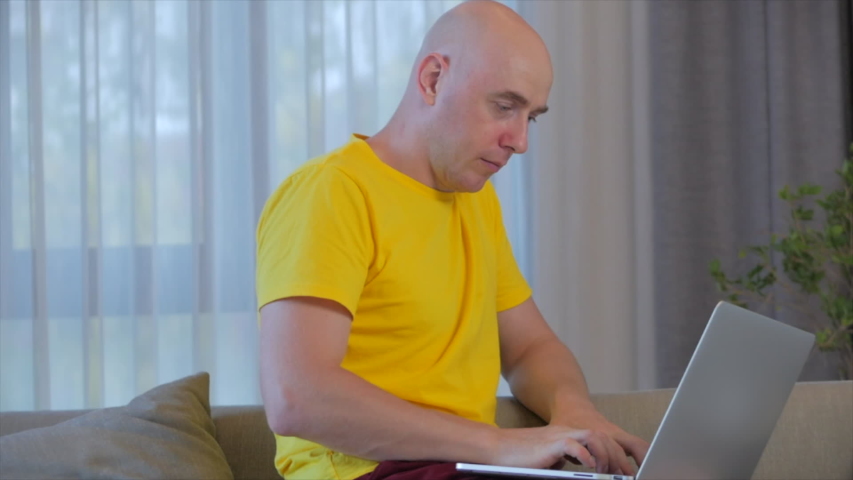 Young serious man freelancer working on a laptop sitting on a sofa at home, businessman typing on a laptop, focused boy using a computer to study online work at home. Home clerk work concept