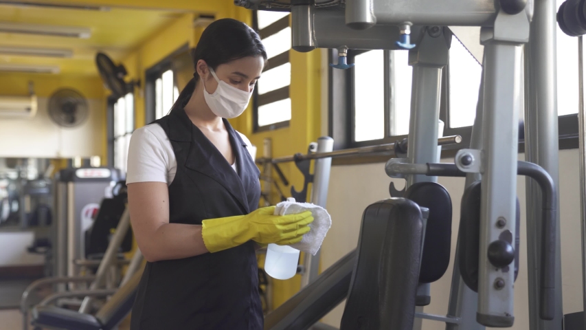 Woman worker disinfects gym fitness equipment from coronavirus covid-19 with antibacterial sanitizer sprayer on quarantine. Cleaner in protective mask cleans training apparatus at workout area.
 | Shutterstock HD Video #1052159173