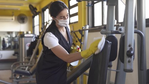 woman worker disinfects gym fitness equipment from coronavirus covid-19 with antibacterial sanitizer sprayer on quarantine. Cleaner in protective mask cleans training apparatus at workout area.
