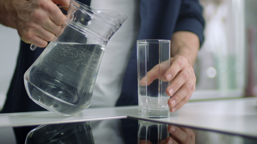 Closeup male hands pouring water into glass from jug at kitchen background. Unrecognizable person drinking water at home kitchen. Unknown man standing in modern home alone. | Shutterstock HD Video #1052159635