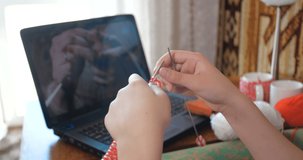 Close up hands of girl. Schoolgirl learning to knit while sitting in a room on couch and watching video lessons from the Internet on a laptop screen. Creative leisure staying at home