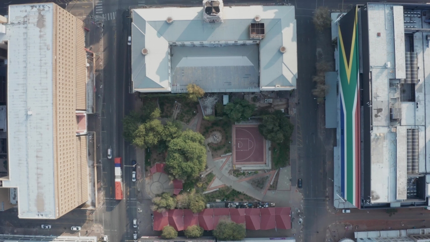 Aerial top view of an inner city park in Johannesburg featuring green trees, a basketball court, and a huge South African flag painted on a building. 