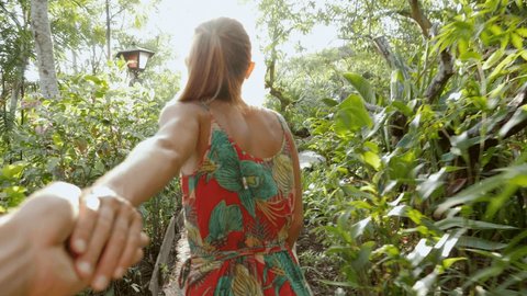 Couple holding hands woman leading the way. Man pov following girlfriend in tropical garden at sunset enjoying nature together. Young couple sharing love smiling and happiness hand in hand 