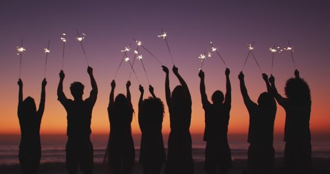 Rear view of a group of friends on holiday enjoying time together on a tropical beach at sundown, holding sparklers up in the air, in slow motion