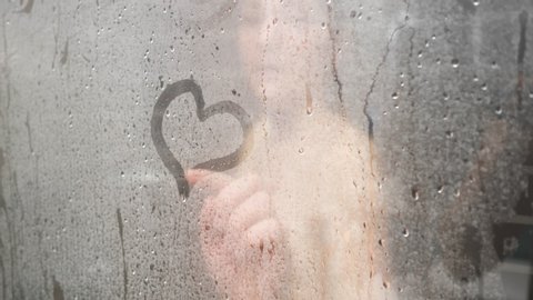 blurry naked young woman hand draws heart on bathroom glass-enclosed shower with white steam at home close view