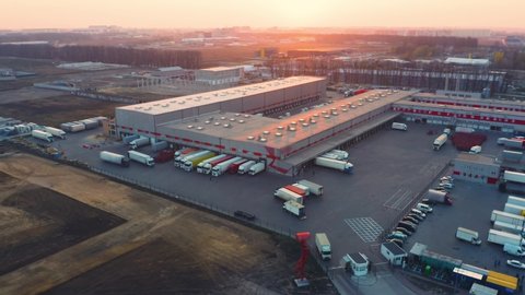 Aerial panoramic view of the logistics park with warehouse, loading hub with many semi trucks with cargo trailers standing at the ramps for loading/unloading goods at sunset