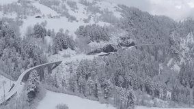 Aerial video of famous train of red color Glacier Express at winter, The train moves to the Landwasser Viaduct and tunnel, landmark of Switzerland, snowing, drone flies forward