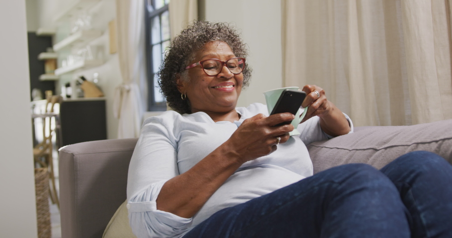 Senior mixed race woman enjoying her time at home, sitting on a couch, drinking tea, using a smartphone, social distancing and self isolation in quarantine lockdown, in slow motion | Shutterstock HD Video #1052209972