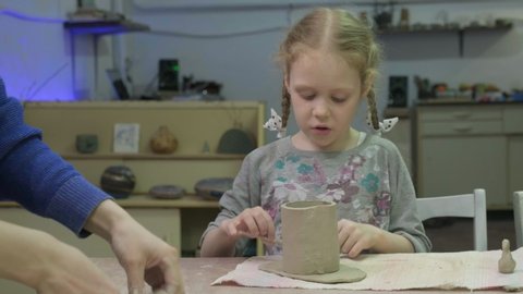Girl preschooler blonde with pigtails with the help of a teacher sculpts a mug of clay. Children's or family clay modeling workshop. Ceramic workshop