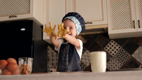 A portrait of a three-year-old girl in a cap and kitchen apron in the kitchen, her face and clothes stained with flour and her hands with dough. The baby shows the camera her hands stained with dough.