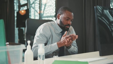 Young african american businessman uses antiseptic for disinfection hands sitting works on the computer coronavirus workplace indoor antibacterial close up slow motion