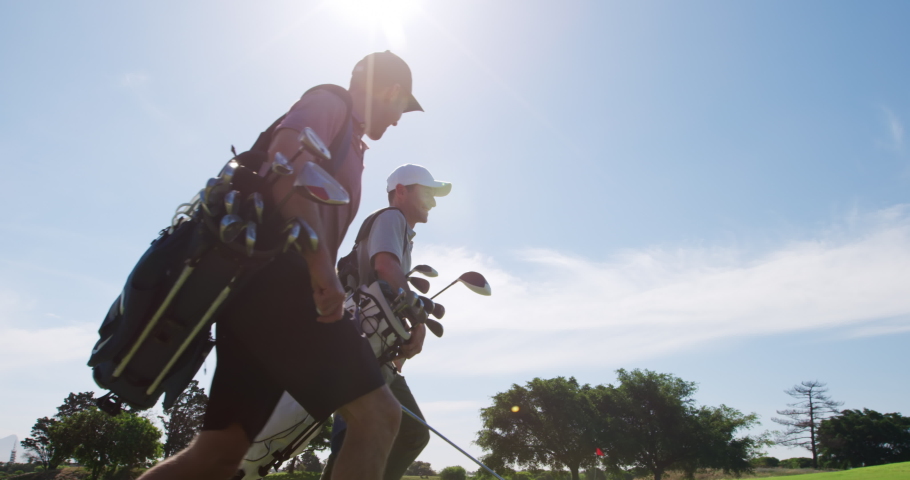 Low angle view of two Caucasian male golfers walking on a golf course on a sunny day wearing caps and golf clothes, holding golf clubs and carrying golf bags, smiling and talking, backlit by sunlight Royalty-Free Stock Footage #1052229739