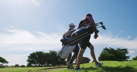 Low angle view of two Caucasian male golfers walking on a golf course on a sunny day wearing caps and golf clothes, holding golf clubs and carrying golf bags, smiling and talking, backlit by sunlight