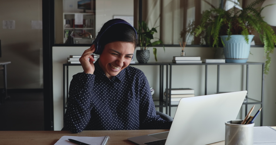 Cheerful young indian business woman wearing headphones laughing at workplace. Funny female office worker having fun listening podcast, video calling, working or studying from home office on laptop. Royalty-Free Stock Footage #1052230138