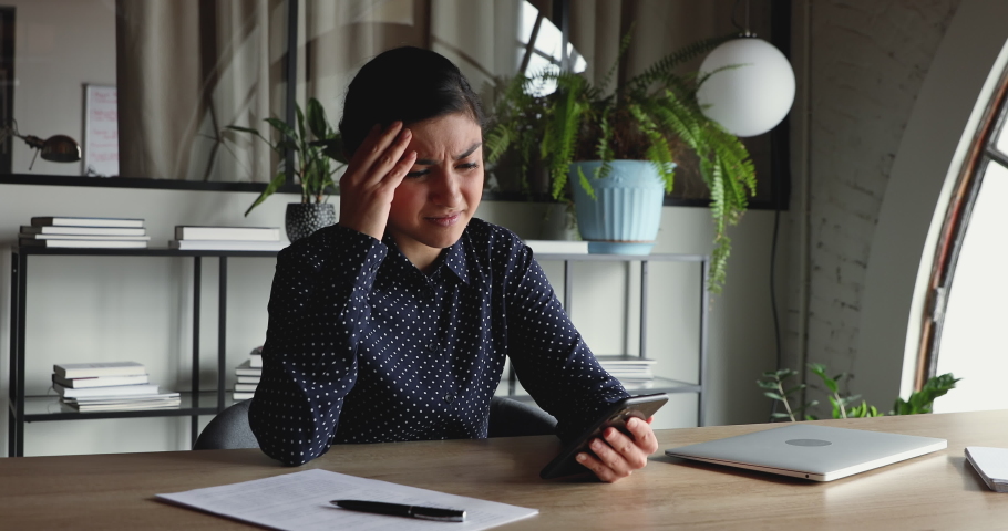 Confused unhappy indian businesswoman feels frustrated about broken phone problem in office. Annoyed female worker reading bad news in message, perplexed by mobile spam sms notification at workplace. Royalty-Free Stock Footage #1052235220